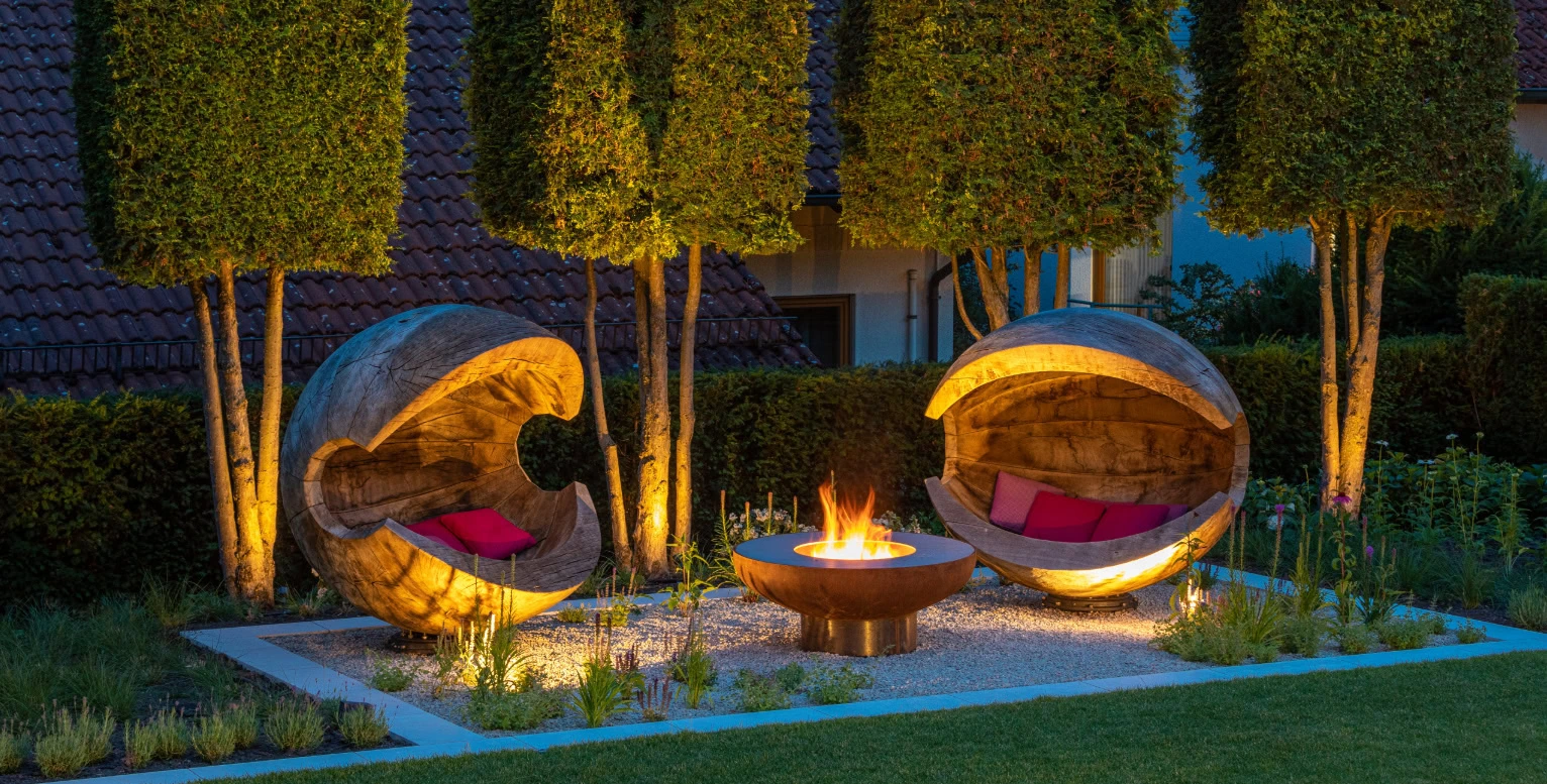 Zwei runde Holz-Sitzgelegenheiten mit Kissen und einer Feuerstelle im Garten bei Nacht.
